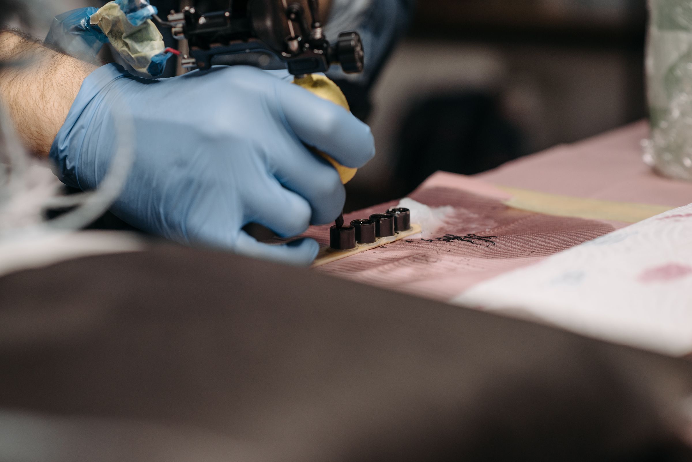 Gloved hand lining up a tattoo machine over cups of black ink
